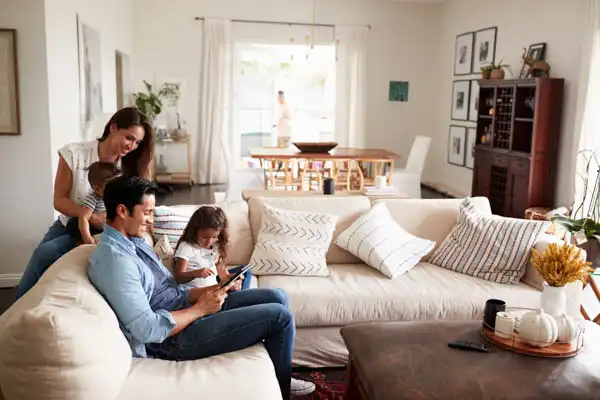 A family sits on a sofa in a bright living room. The father is using a tablet, the daughter is watching, and the mother is smiling. A person is visible in the background near the dining table.