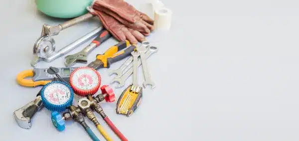 Assorted plumbing tools, ideal for air conditioning repair, including wrenches, pliers, a tubing cutter, gauges, and a pair of gloves, are neatly arranged on a light surface.