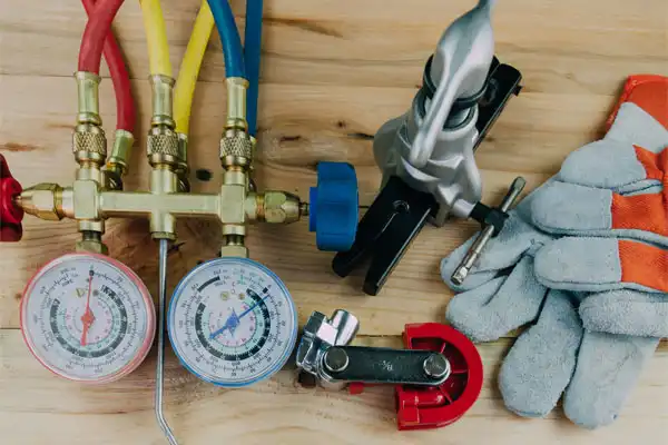 Various HVAC tools on a wooden surface, essential for heating installation, including pressure gauges, red, blue, and yellow hoses, a tube cutter, a flaring tool, and a pair of protective gloves.