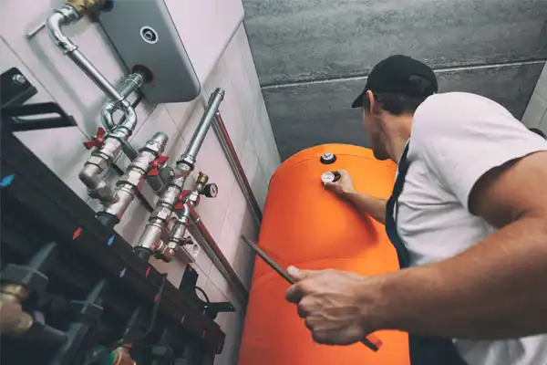 A technician in a cap diligently checks gauges on an orange industrial boiler, surrounded by a maze of pipes and valves, ensuring optimal function for heating repair.