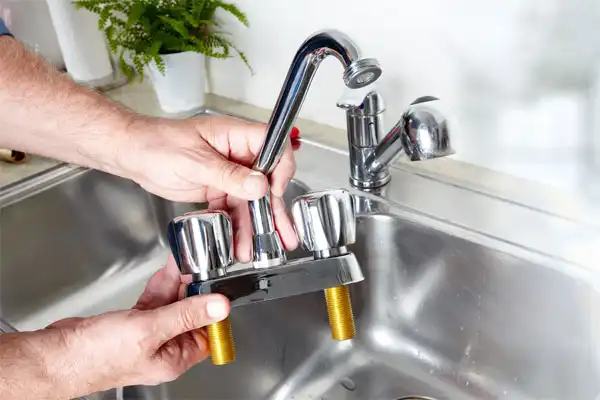 Hands holding a shiny, chrome kitchen faucet with two handles above a stainless steel sink, showcasing expert kitchen plumbing installation.