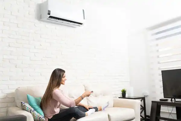 A woman sits on a sofa in her living room, effortlessly using a remote control. Above her, an air conditioner is mounted on the white brick wall, perfectly embodying the benefits of regular mini-split maintenance for efficient cooling.