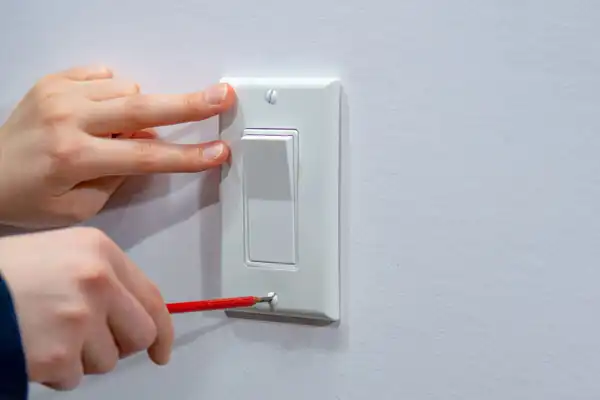 Person using a red screwdriver to install or remove a white light switch plate, with another hand steadying it against the wall.